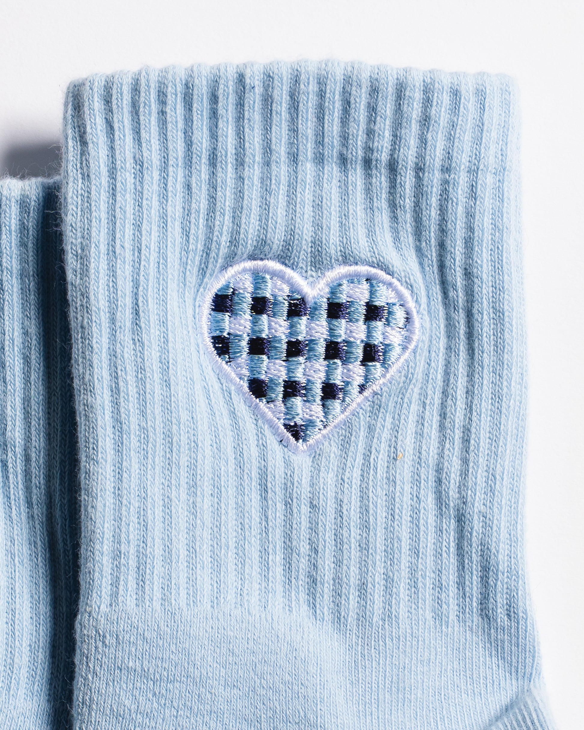 A close-up of The Cross My Heart Grip Sock by Revel Athletic in light blue, showcasing a ribbed design and an embroidered heart patch with a navy blue and light blue checkered pattern inside.