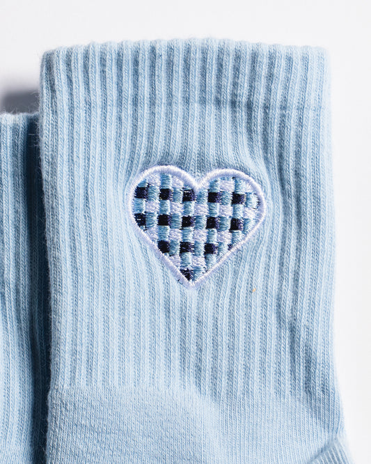 A close-up of The Cross My Heart Grip Sock by Revel Athletic in light blue, showcasing a ribbed design and an embroidered heart patch with a navy blue and light blue checkered pattern inside.