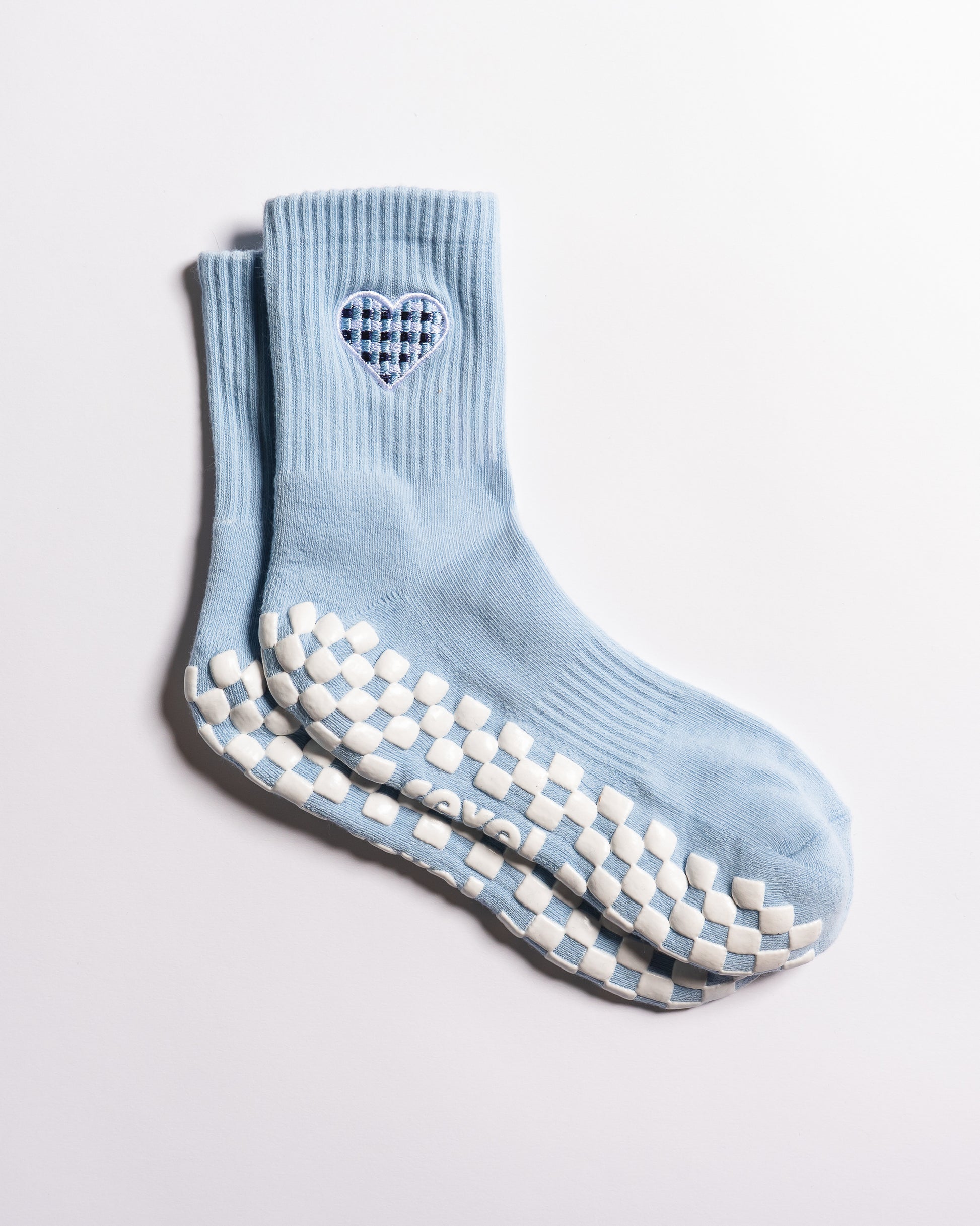 The Cross My Heart Grip Sock by Revel Athletic is shown on a white background, featuring a light blue color with a checkered heart design near the top and a checkered white and blue anti-slip pattern on the soles.