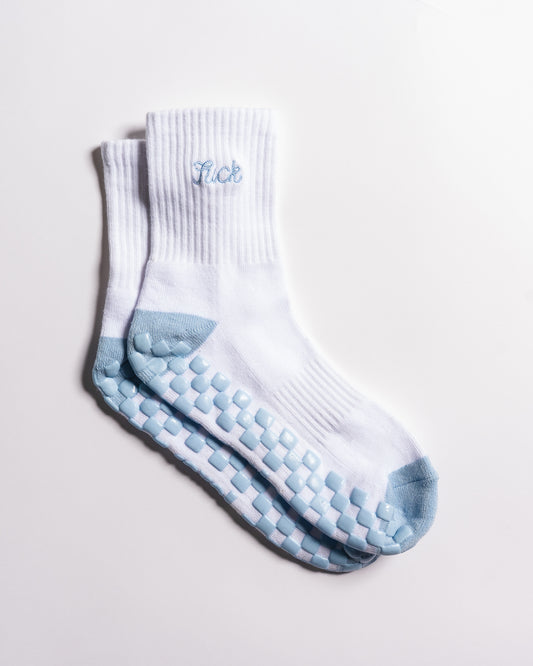 A pair of white Revel Athletic The Pardon My French Grip Socks (Baby Blue) with light blue toes and soles, featuring rubber grip dots for yoga, and the word "lick" embroidered in baby blue near the top.