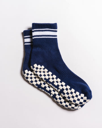 The Revel Athletic “The Country Club Grip Sock” features a blue crew design with two white stripes near the top and white checkered non-slip grips on the soles, displayed against a plain white background. Ideal as barre or Pilates socks, they provide added stability during your workout.