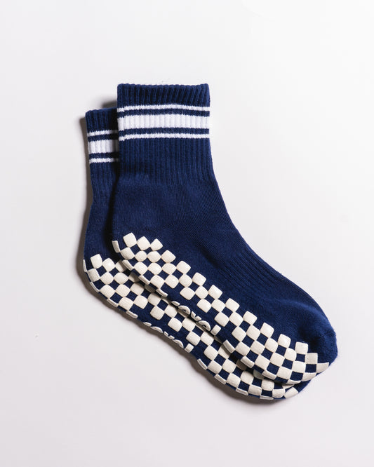 A pair of The Country Club Grip Sock (Navy) by Revel Athletic, featuring navy blue fabric with white stripes at the top and a white and navy checkered non-slip pattern on the soles, displayed on a plain white background.