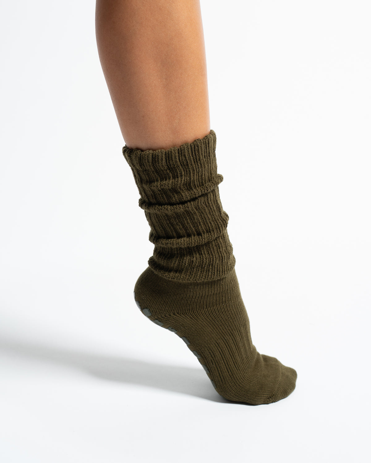 A person’s lower leg and foot wearing Revel Athletic’s “The Slouch - Olive” grip sock, standing against a plain white background.