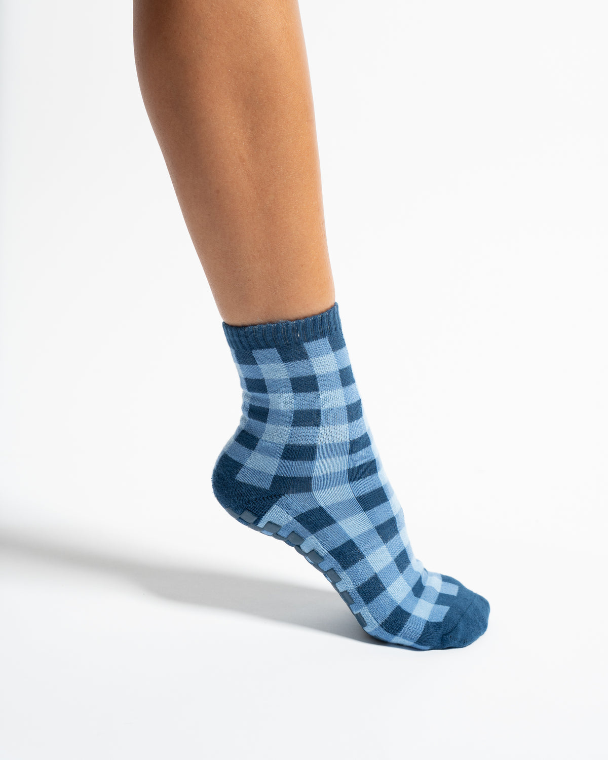 A person’s lower leg and foot wearing a pair of Revel Athletic The Prep Grip Sock in blue gingham, shown against a plain white background.
