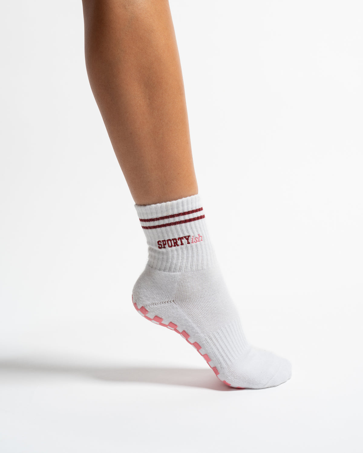 A white Revel Athletic Sportyish crew sock on a foot, featuring two maroon stripes at the cuff, the word SPORTYish in maroon text, and pink grip patterns on the sole.
