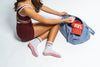 A person in maroon athletic wear and pink checkered pilates grip socks packs their The Sportyish gym bag with a red baseball cap and a small water bottle.