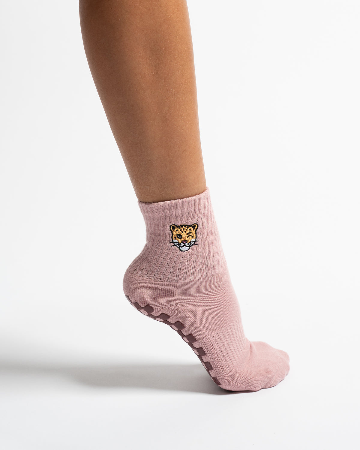 A person wearing The Wildcat Grip Sock by Revel Athletic—featuring a light pink color, a small embroidered cat face, and non-slip grips on the sole—standing on a white background.