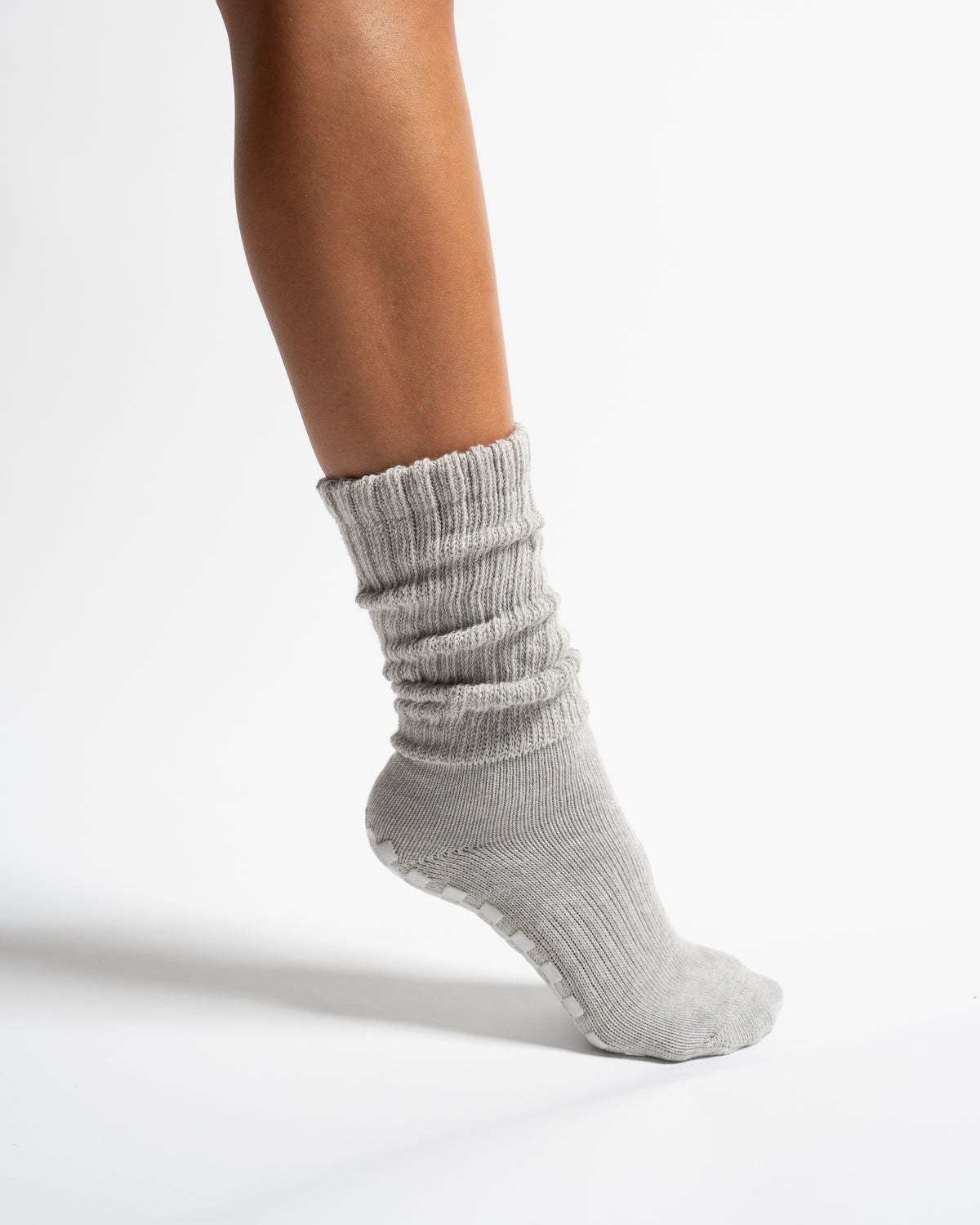 A person’s lower leg wearing Revel Athletic’s The Slouch - Grey Pilates grip sock, featuring a scrunched design and textured grips on the sole, set against a plain white background.