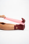 A person sits with feet flexed, stretching The Sweetheart pink resistance band from Revel Athletic with both hands, while wearing maroon crew length socks featuring a yellow emblem, all set against a white background.
