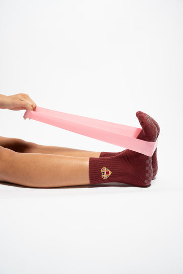 A person sits with feet flexed, stretching The Sweetheart pink resistance band from Revel Athletic with both hands, while wearing maroon crew length socks featuring a yellow emblem, all set against a white background.