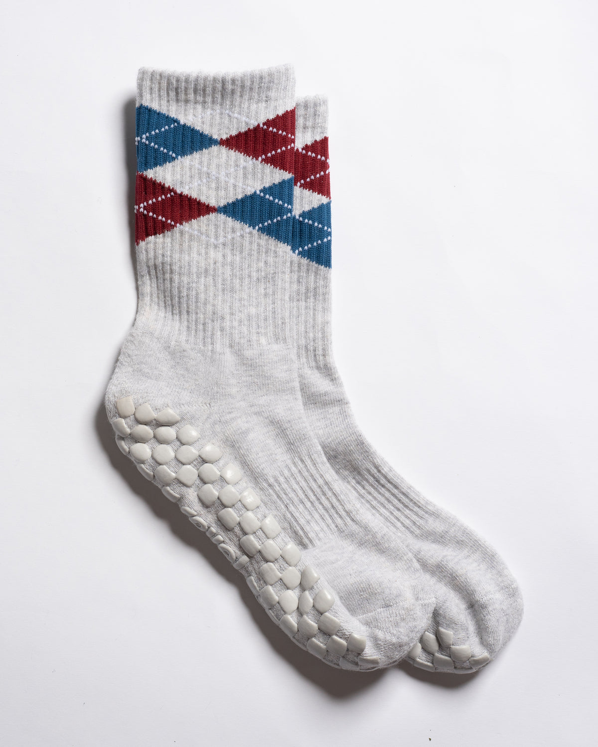 Revel Athletic’s The Legacy socks are white, crew length, featuring red and blue argyle patterns near the top and textured white GripGrid grips on the soles, perfect for Pilates, shown on a white background.