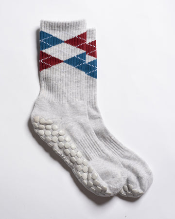Revel Athletic’s The Legacy socks are white, crew length, featuring red and blue argyle patterns near the top and textured white GripGrid grips on the soles, perfect for Pilates, shown on a white background.