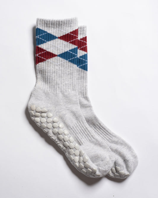Revel Athletic’s The Legacy socks are white, crew length, featuring red and blue argyle patterns near the top and textured white GripGrid grips on the soles, perfect for Pilates, shown on a white background.