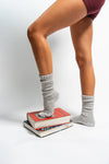 A person wearing The Slouch - Grey grip socks by Revel Athletic and maroon shorts stands with one foot on a stack of books and the other on the floor, set against a white background.