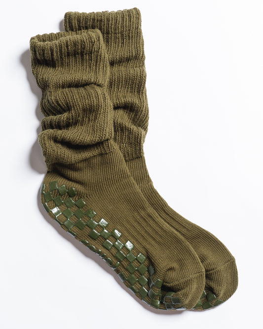 A pair of Revel Athletic’s The Slouch - Olive knit Pilates socks, featuring a relaxed fit in olive green with dark green checkerboard-patterned non-slip grips on the soles, displayed against a white background.