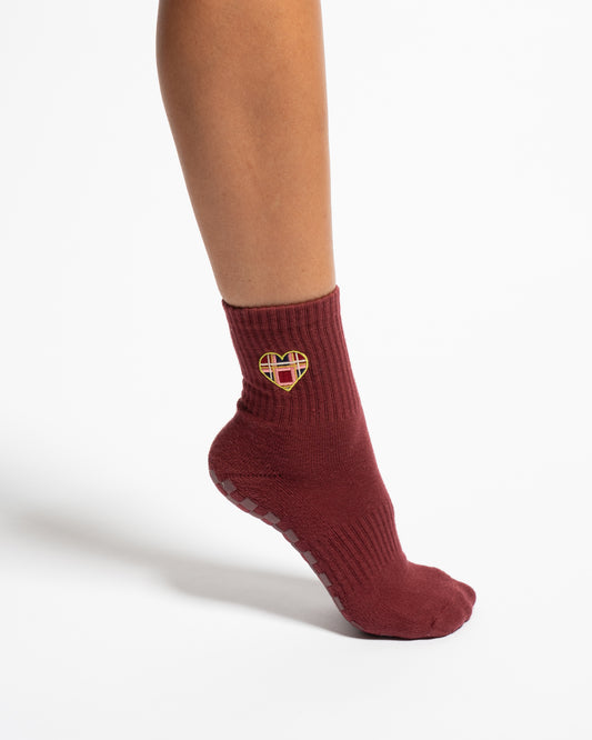 A person wears The Sweetheart by Revel Athletic—a maroon crew-length sock adorned with a plaid heart-shaped patch. Set against a white background, this stylish sock is perfect for Pilates sessions thanks to its secure GripGrid sole.