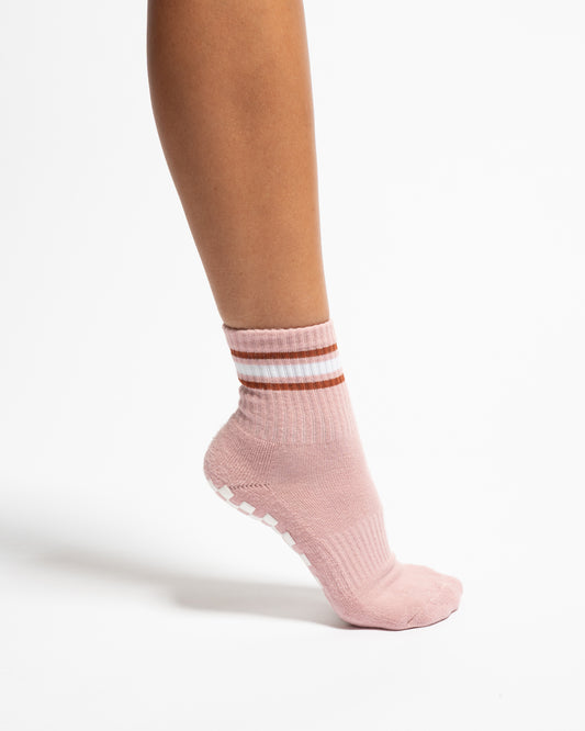 The Rally by Revel Athletic is a light pink, ankle-length Pilates sock featuring two horizontal brown and white stripes at the cuff and white grip pads on the sole, styled with a retro court aesthetic.