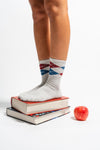 A person wearing Revel Athletic’s The Legacy crew length patterned grip socks stands on two stacked books, with a red apple placed beside the books on a white background—perfect for adding balance and comfort to your Pilates or daily routine.