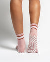 Back view of legs wearing The Rally retro court style pink crew socks by Revel Athletic, featuring white and brown striped cuffs and white checkered silicone grips on the soles.