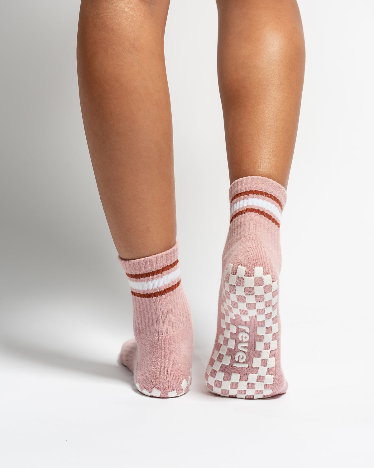 Back view of legs wearing The Rally retro court style pink crew socks by Revel Athletic, featuring white and brown striped cuffs and white checkered silicone grips on the soles.