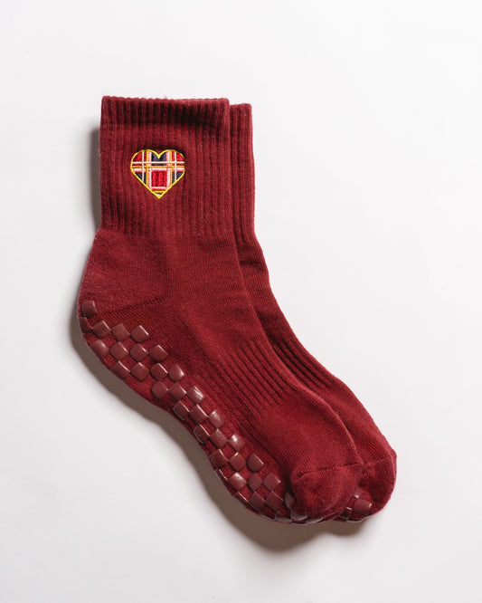 A pair of maroon crew length socks from Revel Athletic, called The Sweetheart, featuring GripGrid rubber soles for Pilates and a heart-shaped patch with a yellow, red, and white pattern near the top.