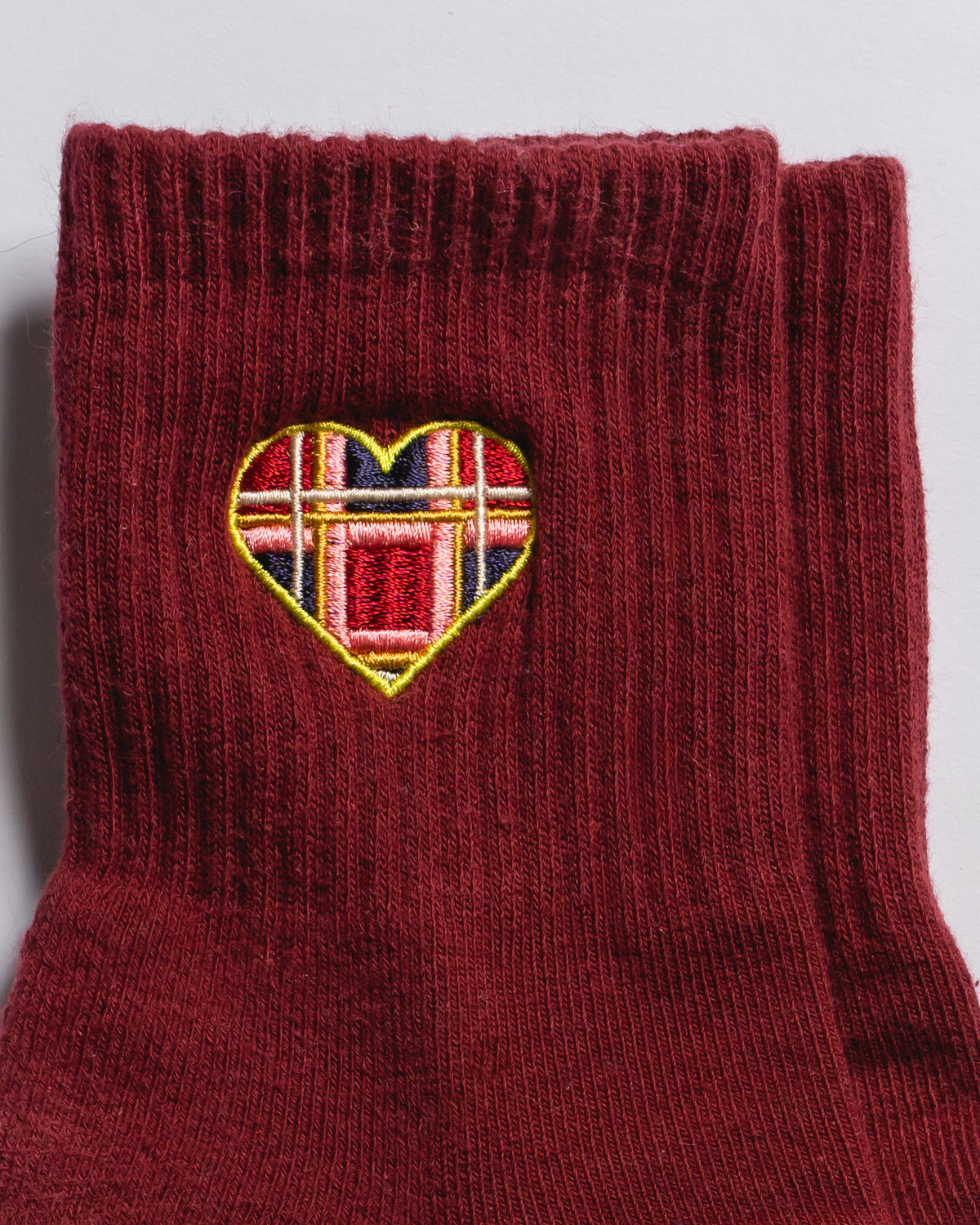 The Sweetheart by Revel Athletic are maroon ribbed crew socks featuring a small heart-shaped patch with a red, blue, yellow, and white plaid embroidery near the top—perfect for Pilates or studio workouts.
