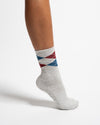 A person’s lower leg and foot wearing Revel Athletic’s The Legacy gray crew length socks with blue and red diamond patterns, featuring GripGrid soles ideal for Pilates, shown against a plain white background.
