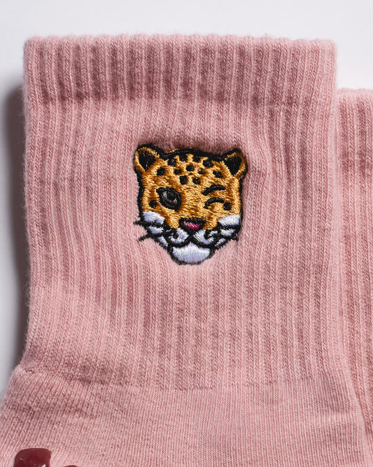 Step into style with The Wildcat Grip Sock by Revel Athletic—pink ribbed socks showcasing a winking, embroidered yellow and black leopard face with white detailing and dark spots, ideal for pilates or relaxed lounging.