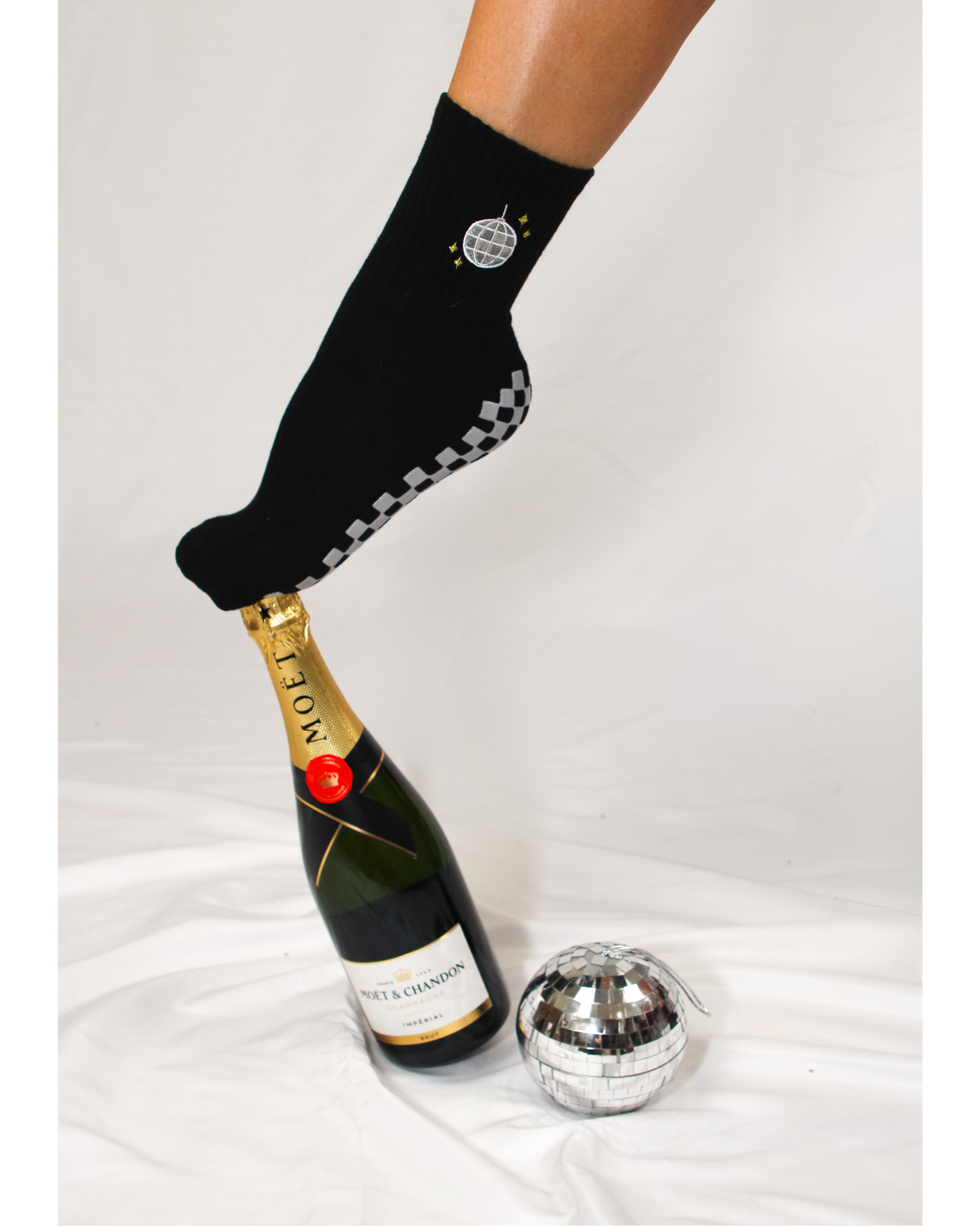A foot in The All Nighter Grip Sock by Revel Athletic—featuring a black sock with a disco ball design—balances on a capped Moët & Chandon champagne bottle, with a small disco ball nearby on a white fabric background, making it the perfect accessory to elevate your Pilates or yoga style.