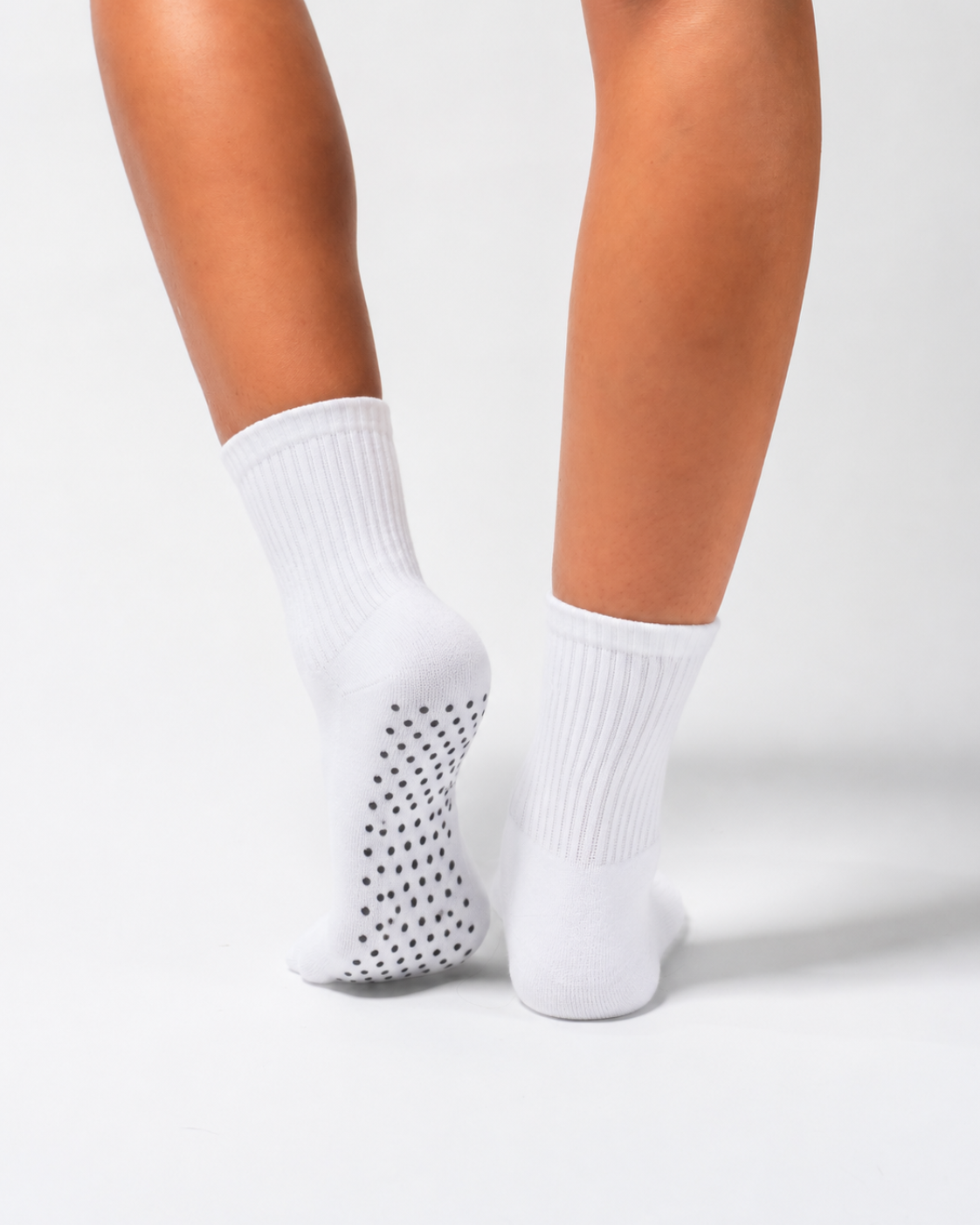 A person wearing white crew pilates socks with black non-slip grips on the bottom, standing on a white surface with their heels raised.